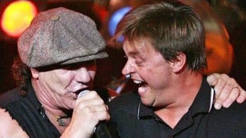 Brian Johnson of AC/DC and comedian Jim Breuer singing together on stage, sharing a microphone and smiling during a their AC/DC jam.