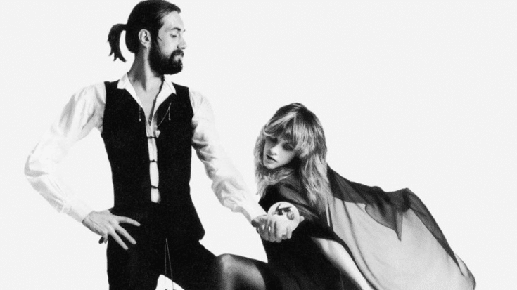 Black-and-white promotional photograph of Mick Fleetwood and Stevie Nicks of Fleetwood Mac from their photoshoot for Rumours. Mick Fleetwood stands in a theatrical pose wearing a vest over a white shirt, while Stevie Nicks, dressed in a flowing black dress with sheer fabric, gracefully holds his hand.