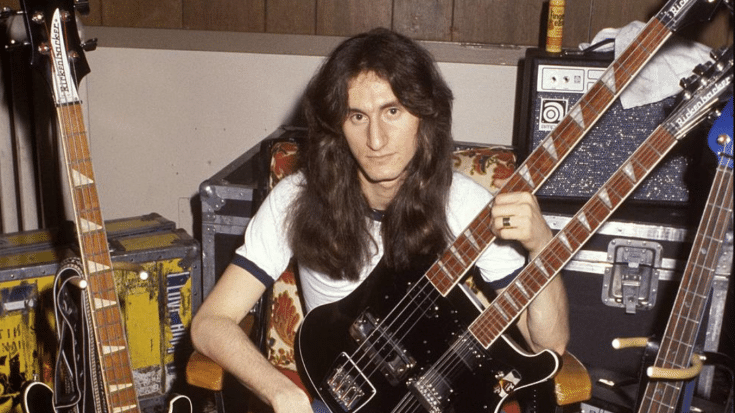 Geddy Lee of Rush sitting backstage, holding a black double-neck Rickenbacker bass guitar. Surrounded by various bass guitars, amplifiers, and equipment cases.