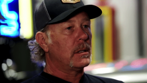 James Hetfield speaking about his custom car collection during an interview, wearing a black cap and shirt, with classic cars blurred in the background.