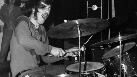 Black and white photo of John Bonham playing the drums with intense energy. He is mid-performance, gripping the drumsticks tightly, with his mouth open and hair flying as he strikes the drums.