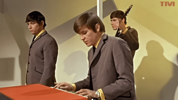 A colorized still of three musicians from the 1960s British band The Animals performing on a television stage. The keyboardist in the foreground is focused on playing, while two other band members stand behind him, one holding a guitar.