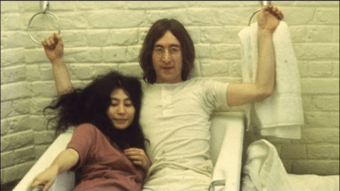 John Lennon and Yoko Ono sitting together in a white bathtub against a brick wall.
