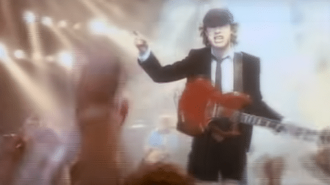 Angus Young of AC/DC performing on stage in his signature schoolboy uniform, playing a red Gibson SG guitar.