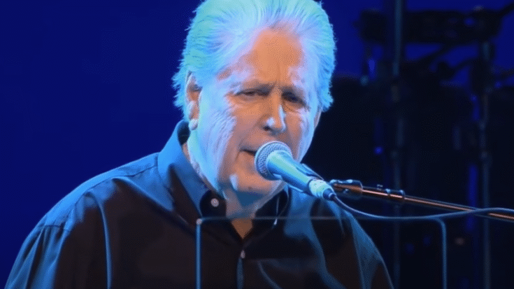 Brian Wilson performing live on stage, singing into a microphone under blue lighting.