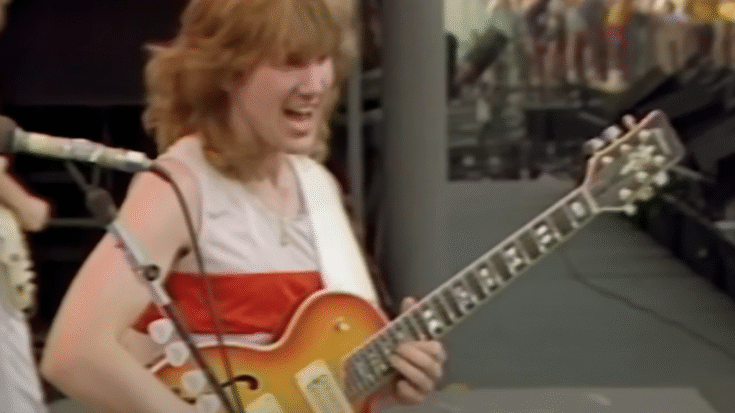 Rik Emmett of Triumph performing live at the US Festival 1983, smiling while playing a sunburst electric guitar on stage.