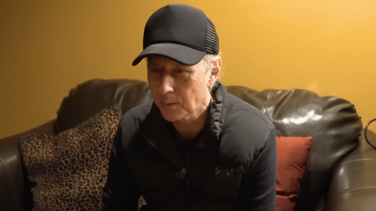 Jerry Cantrell sitting on a leather couch during an interview with Rock Feed, wearing a black cap and jacket.