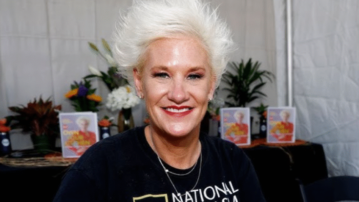 Anne Burrell smiling at an event while seated in front of a table decorated with flowers and copies of her cookbook