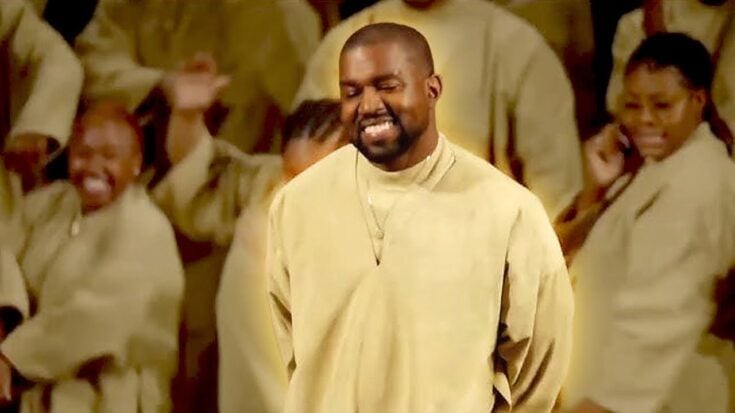 Kanye West smiling during a live Sunday Service performance, dressed in beige robes with choir members in the background.