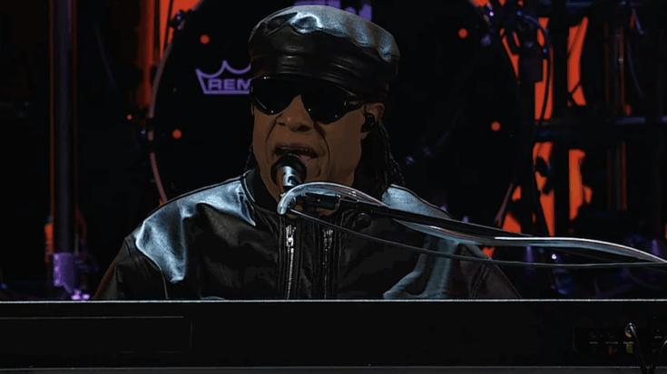 Stevie Wonder performing live in a black leather outfit and sunglasses, playing the keyboard on stage under dramatic lighting.