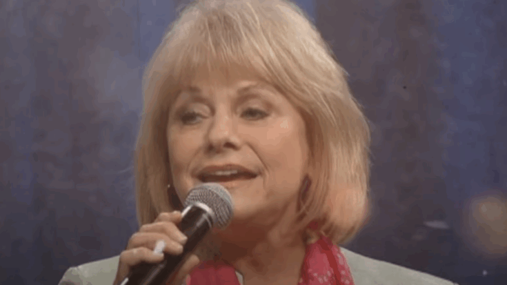 Helen Cornelius performing on stage with a microphone, wearing a light blazer and pink scarf.