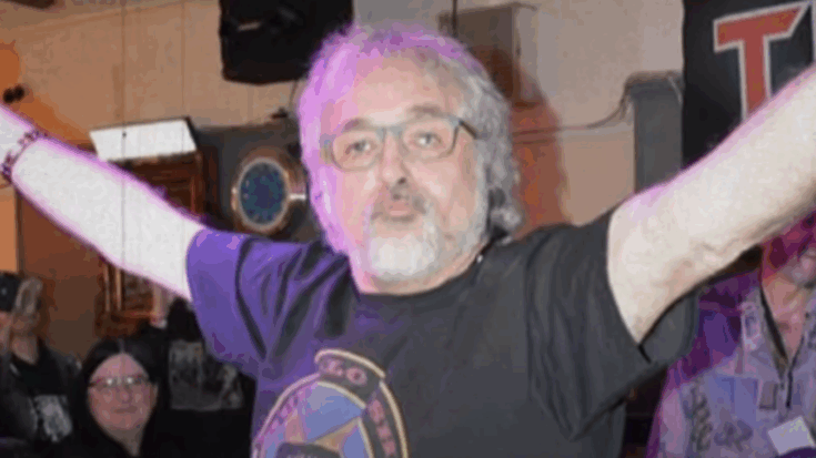 Paul Mario Day, Iron Maiden’s original vocalist, with arms raised during a fan event — smiling and wearing a vintage rock band shirt.