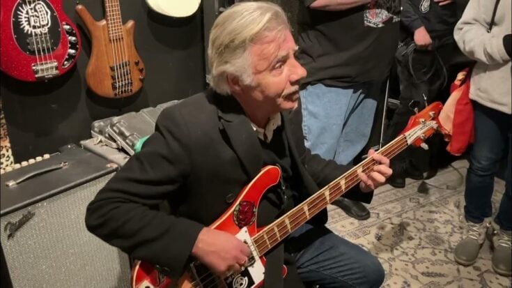 Glen Matlock playing a red bass guitar at an intimate event, surrounded by fans and guitars on display.