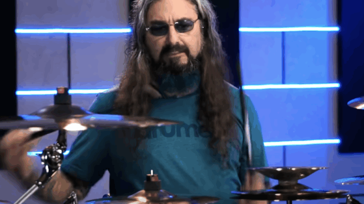 Mike Portnoy playing drums in a studio, wearing sunglasses and a blue shirt, with cymbals in motion.