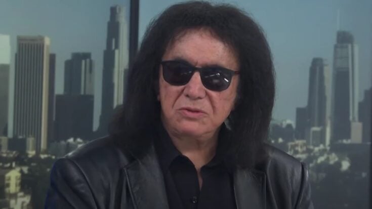 Gene Simmons wearing sunglasses and a leather jacket during a television interview, with a city skyline in the background.