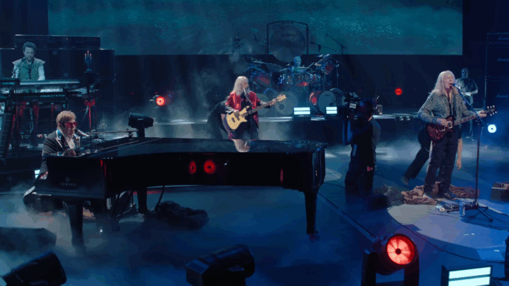 Spinal Tap performing live on stage with Elton John at the piano, surrounded by fog, dramatic lighting, and a full rock band setup.