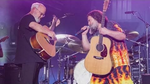 Jack Black and Kyle Gass of Tenacious D performing live on stage, both playing acoustic guitars during a high-energy concert.