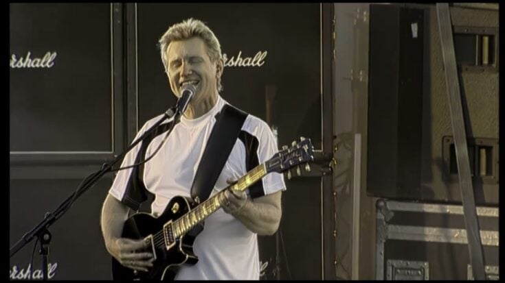 Rik Emmett of Triumph performing live on stage with a guitar and microphone, backed by Marshall amplifiers.