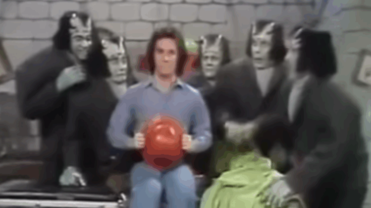 A 1970s Saturday morning TV scene featuring a man holding a red helmet, surrounded by people dressed as Frankenstein-style monsters in a playful set.