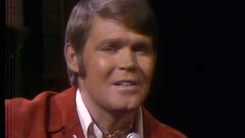 Glen Campbell performing on stage in the late 1960s, wearing a red jacket and singing into a microphone.