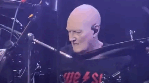 Chris Slade performing energetically on stage, focused behind his drum kit under concert lighting.