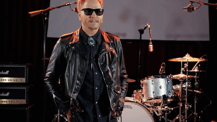 Matt Sorum wearing a leather jacket and sunglasses while standing in front of a drum set on stage.