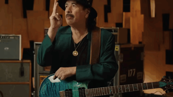 Carlos Santana gestures while holding his guitar during a studio session, surrounded by amplifiers and equipment cases.