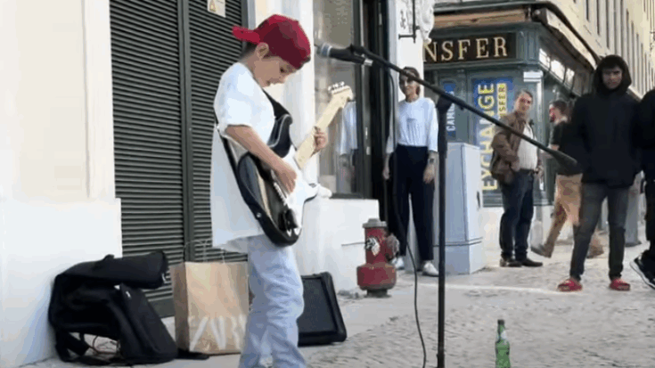Eight-year-old guitarist Leonardo Patrick performs an impressive street cover of Bronski Beat’s “Smalltown Boy” at a busy outdoor mall, captivating onlookers with his electric guitar skills and looping setup.
