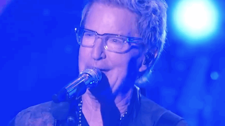 Kevin Cronin performs live on stage, singing into a microphone under blue stage lights during a recent REO Speedwagon appearance.