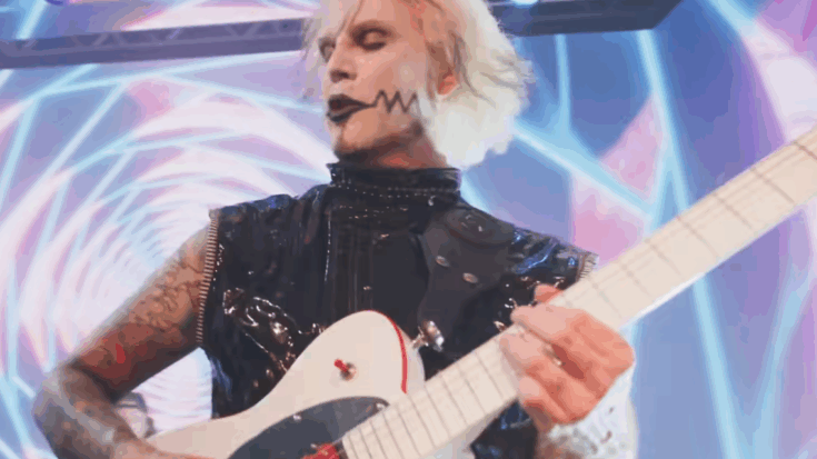 John 5 performing on stage with his signature white Telecaster guitar featuring red pickups, wearing black stage attire under colorful lights.
