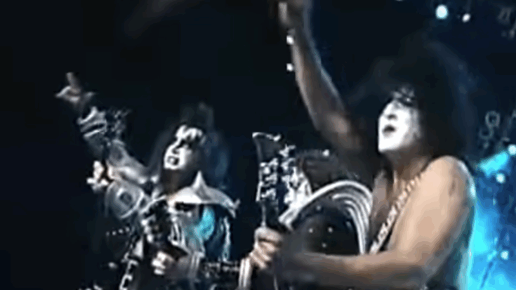 KISS performing live onstage in full makeup and costumes, with Gene Simmons and Paul Stanley playing guitars under bright stage lights.
