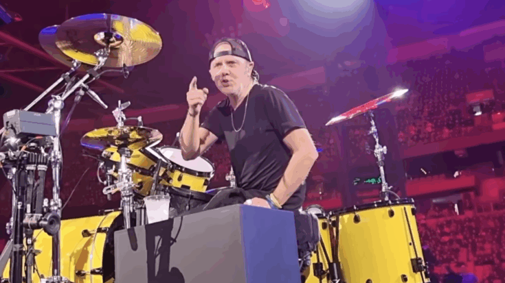 Lars Ulrich performing on stage behind his yellow drum kit during a Metallica concert, wearing a backward cap and gesturing to the crowd.