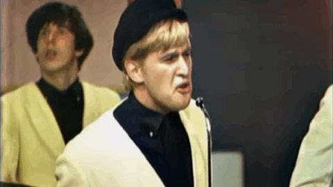 Jack Ely performing “Louie Louie” with The Kingsmen in 1963, wearing a light jacket and beret while singing into a microphone during a live television appearance.