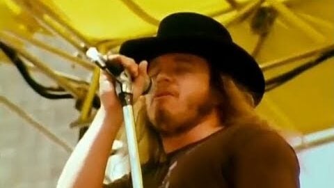 Ronnie Van Zant performing live on stage, wearing a black hat and singing into a microphone under a yellow canopy.