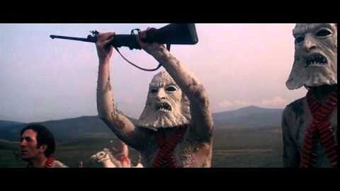 Masked figures wearing primitive costumes and ammunition belts raise rifles in a dramatic scene from the 1974 sci-fi film Zardoz, set against a desolate landscape.