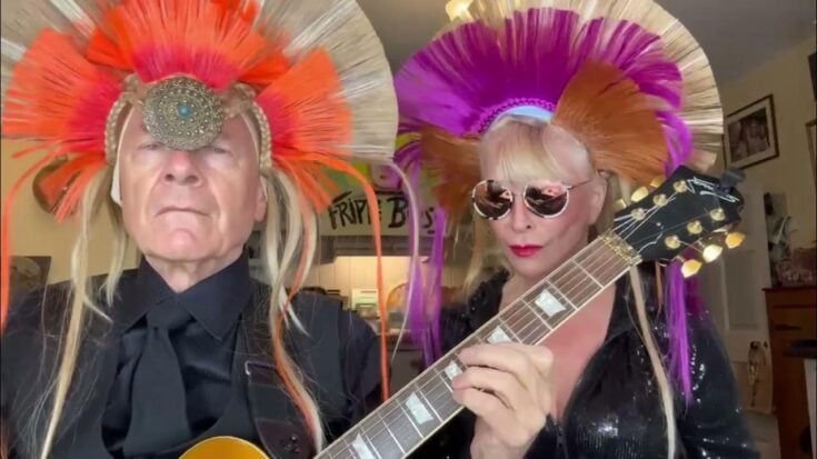 Toyah and Robert Fripp wearing colorful feathered headdresses play guitar together during a lively at-home performance.