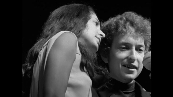 A black-and-white photo of Joan Baez and Bob Dylan performing closely together on stage during the 1960s folk era.