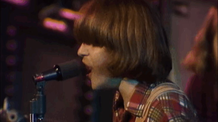 John Fogerty performing live at The Royal Albert Hall with Creedence Clearwater Revival, singing into a microphone while wearing a plaid shirt, captured during a classic rock performance with warm stage lighting.