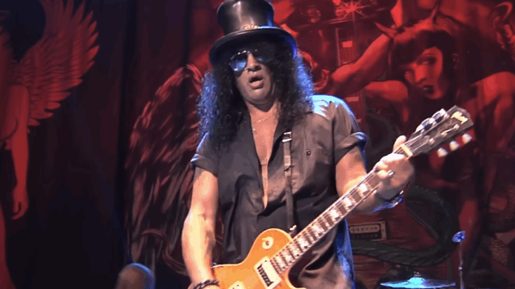 Slash performing live on stage with his Les Paul guitar and signature top hat, captured during a concert performance.