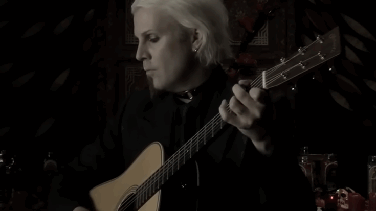 John 5 plays an acoustic guitar in a dimly lit room, creating an intimate and reflective scene.