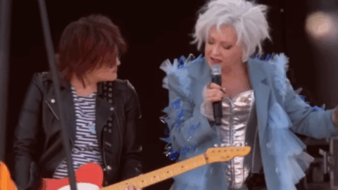 Cyndi Lauper performing on stage with a guitarist during a live concert, wearing a silver outfit and blue ruffled jacket.
