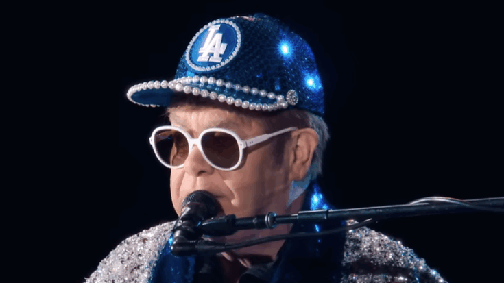 Elton John wearing a sequined blue Dodgers cap and white sunglasses playing piano onstage during a live performance.