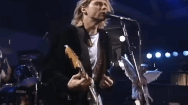 Kurt Cobain performing live onstage, singing into a microphone while playing an electric guitar during a concert.