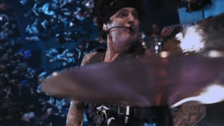 Tommy Lee performing on a drum kit during a live show, wearing a headset microphone with stage lights in the background.