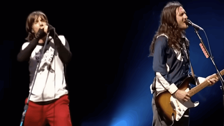 Two members of the Red Hot Chili Peppers performing onstage under concert lighting, with one singing into a microphone and the other playing guitar.