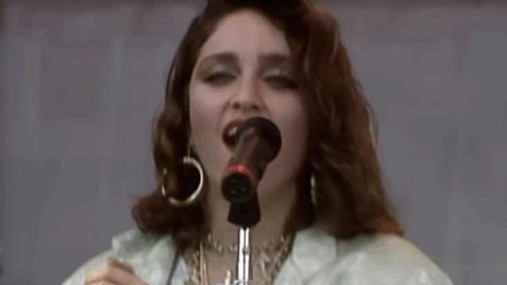 Madonna singing on stage at the 1985 Live Aid concert, wearing large hoop earrings and layered jewelry while performing in front of a live audience.