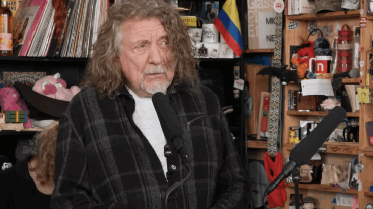 Robert Plant performing with Saving Grace during his NPR Tiny Desk Concert, singing into a microphone in the intimate, cluttered office-style setting.