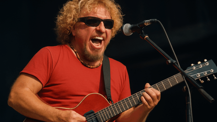 Sammy Hagar performing live onstage, wearing sunglasses and a red shirt while playing guitar and singing into a microphone.