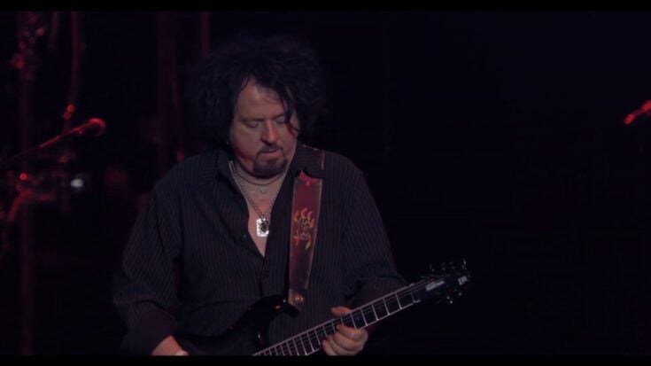 Steve Lukather is performing live onstage, playing an electric guitar under dramatic lighting during a concert.
