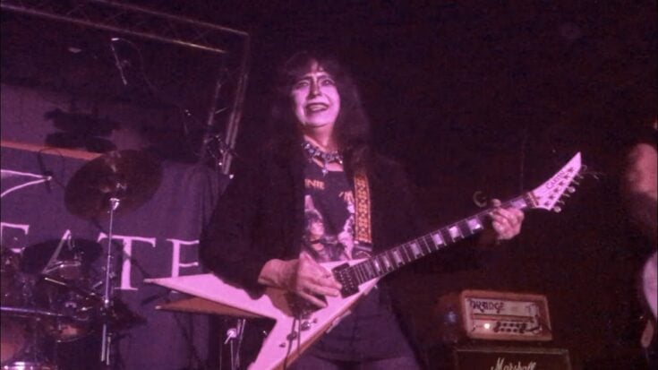 Vinnie Vincent performing on stage with his signature Flying V guitar, wearing makeup and an androgynous glam rock outfit under purple stage lighting.